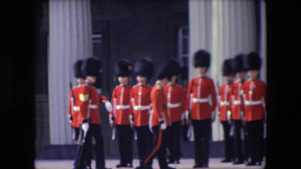 1967 London home-movie still showing ceremonial guards in red tunics and dark hats clustered in formation beside a pale column.