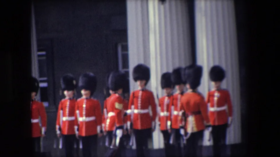 1967 London frame showing a wider group of ceremonial guards in red uniforms and tall dark hats standing near large columns and a dark building wall.