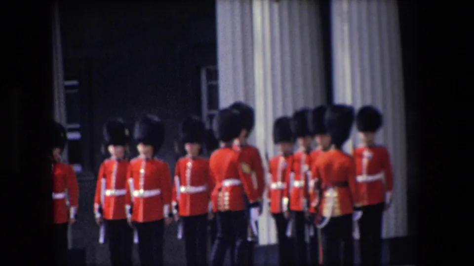 Royal Guard soldiers in red tunics and dark ceremonial hats stand in formation near pale architectural curtains or columns in London, supported by the 1967 Buckingham Palace metadata.