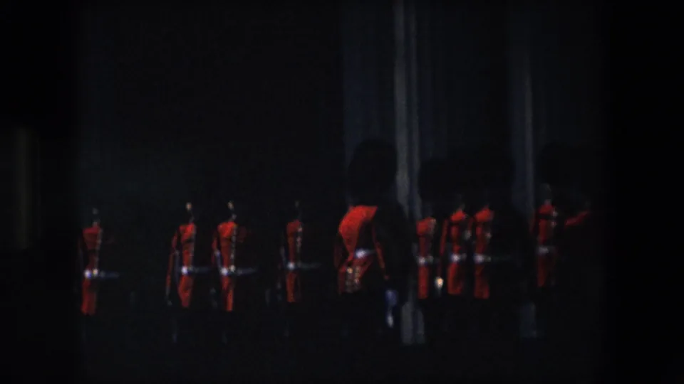 A line of Royal Guard soldiers in red uniforms and dark hats appears in a dimly exposed ceremonial area, with one closer figure partly visible at right.
