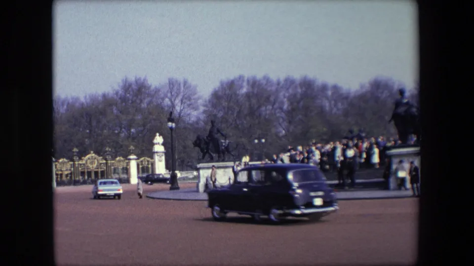 Period cars travel across the roadway outside Buckingham Palace gates, with pedestrians, statues, trees, and a crowd visible in the background.