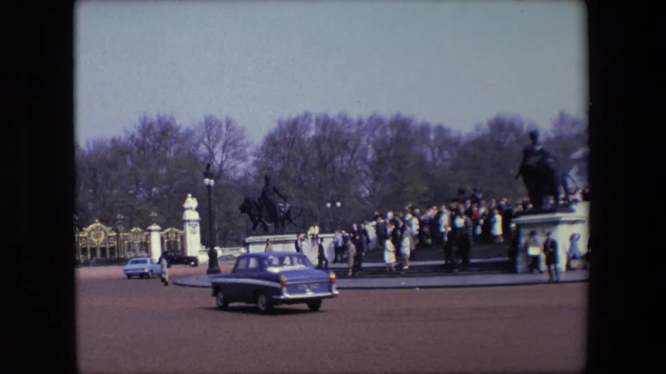 A mid-century car crosses the foreground outside Buckingham Palace gates while pedestrians cluster near statues and trees in the distance.