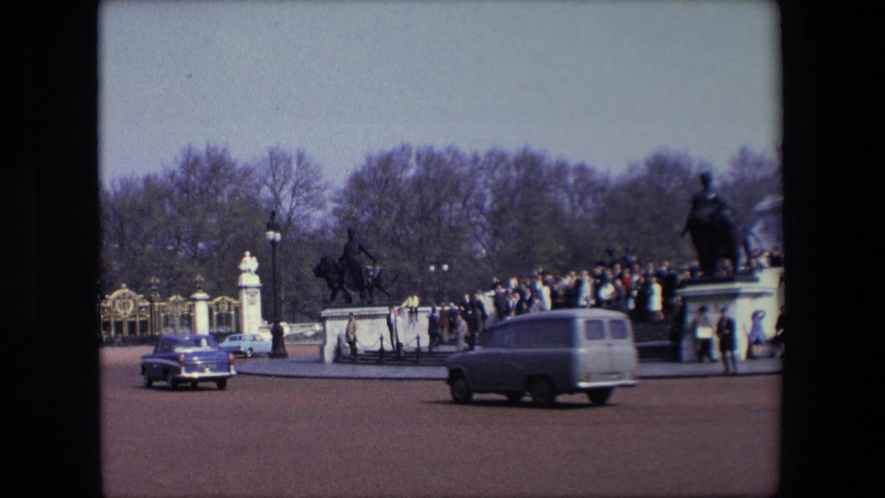 Classic cars pass a broad road in front of gates, pedestrians, and a crowd near a monument.