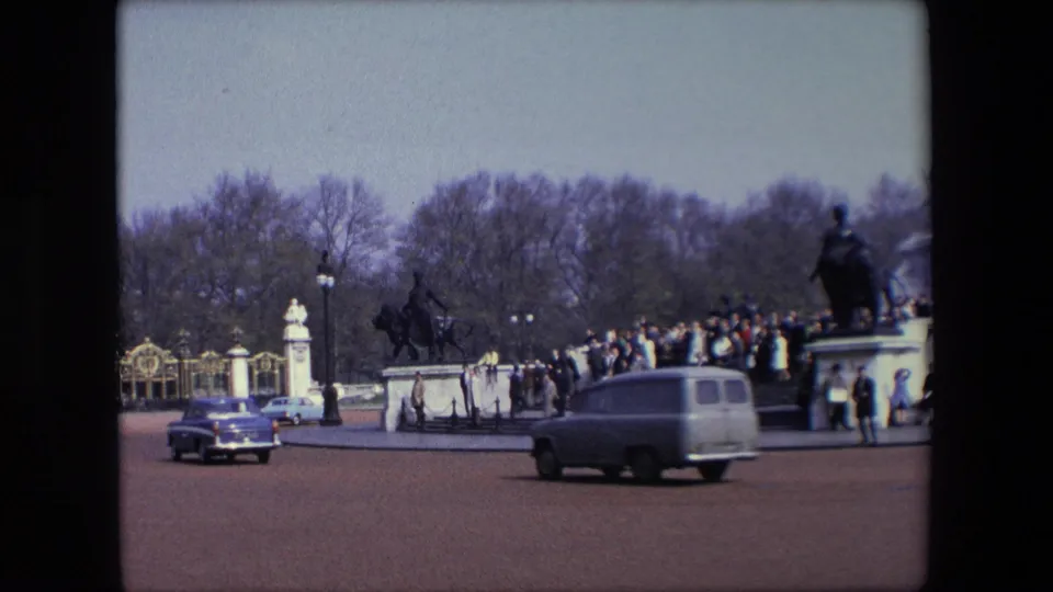 1967 London home-movie frame showing classic cars on a wide street with ornate gates, pedestrians, bare trees, and visitors gathered near a public monument.