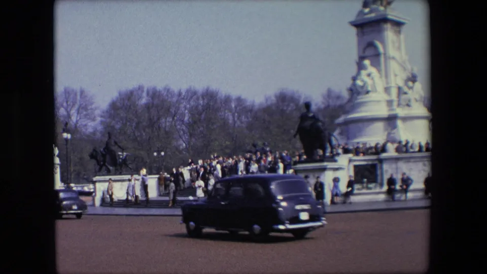 1967 London home-movie still showing a black classic car passing close to camera with pedestrians and a crowd around a large public monument behind it.
