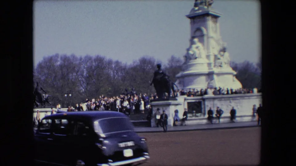 1967 London frame with a dark classic car in the foreground and people gathered around a stone monument and statue in the background.
