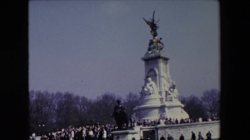 1967 London home-movie frame showing a tall public monument with sculptural figures, a small crowd below, bare trees, and dark 8mm frame edges.