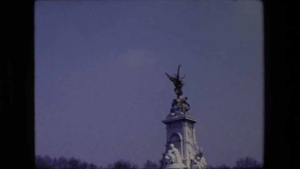 1967 London home-movie frame with the monument set low in the composition, showing its sculptural top, pale stonework, bare tree line, and broad sky.