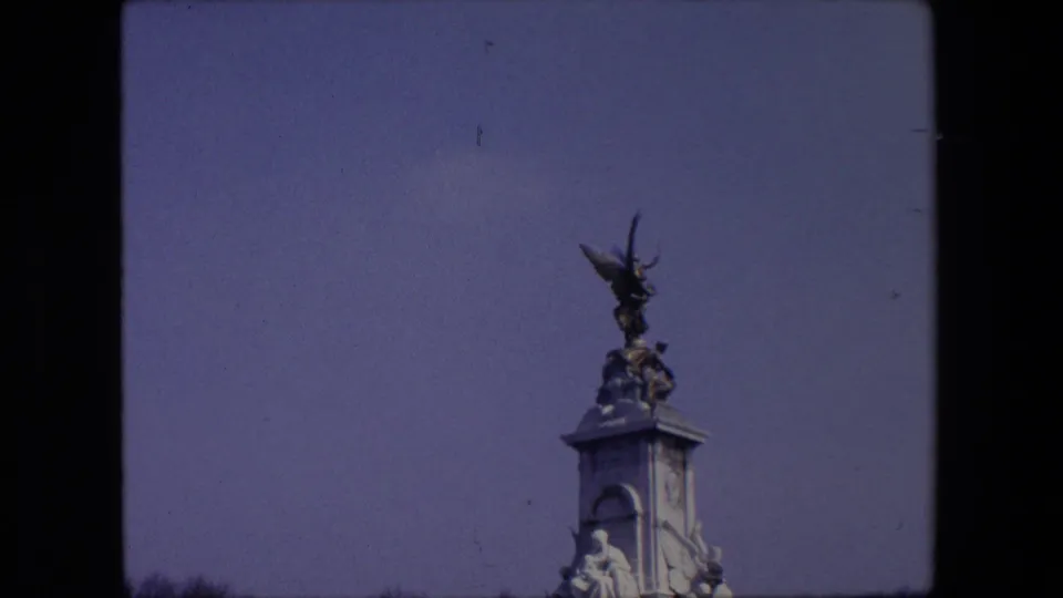 1967 London home-movie frame showing the public monument from farther back, with sculptural figures visible near the top and a dark tree line below.