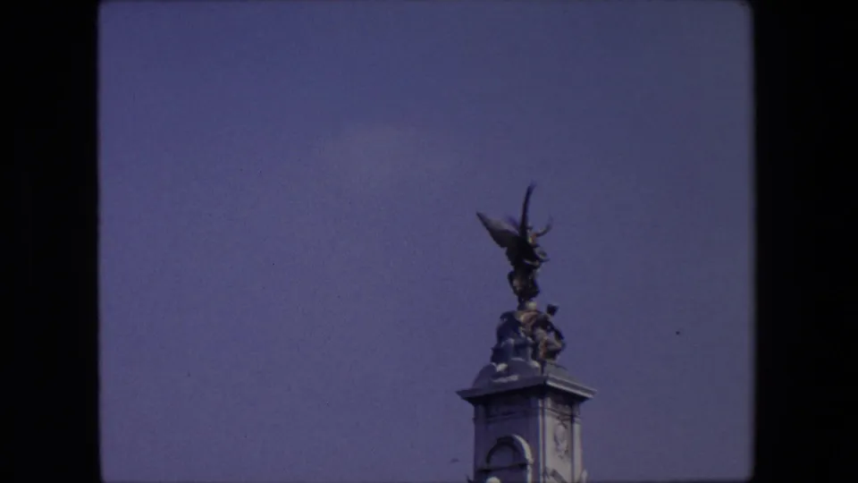 1967 London home-movie frame showing the upper part of a stone pillar with a winged statue and sculptural figures silhouetted against a blue sky.