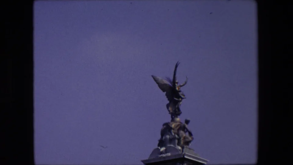 Home-movie view from London in 1967 showing a dark winged statue and clustered figures on the top of a stone monument against a clear blue sky.