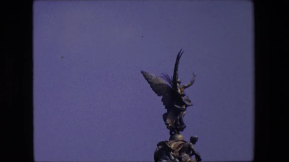Closer 8mm frame of the London monument, with the winged figure and base sculptures filling more of the view against the blue sky.