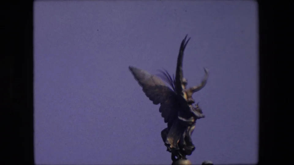 A 1967 London 8mm frame shows a dark winged statue with one arm raised, filmed from below against a blue sky.