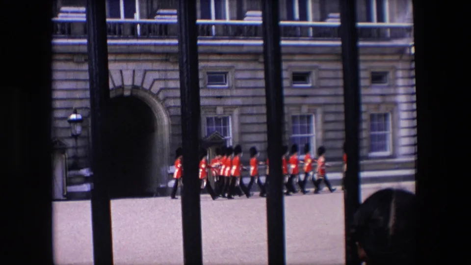 Uniformed guards in red jackets move across the Buckingham Palace forecourt as seen through tall black railings in 1967 London footage.