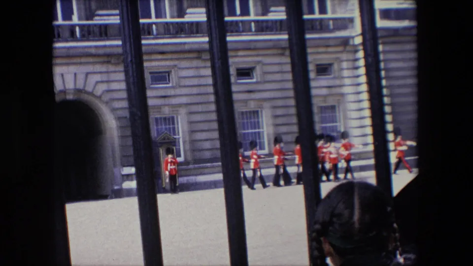 A group of Buckingham Palace guards stands and moves near the facade while a visitor’s head is visible close to the railings in 1967 London home-movie footage.
