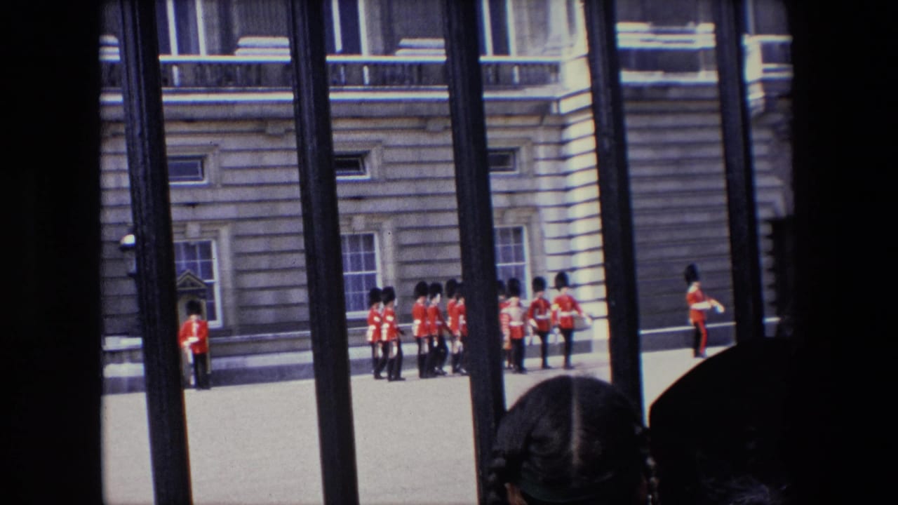 1967 London home-movie frame showing Queen's Guard soldiers in red uniforms near a palace facade, filmed through dark iron railings with spectators partly visible in front.