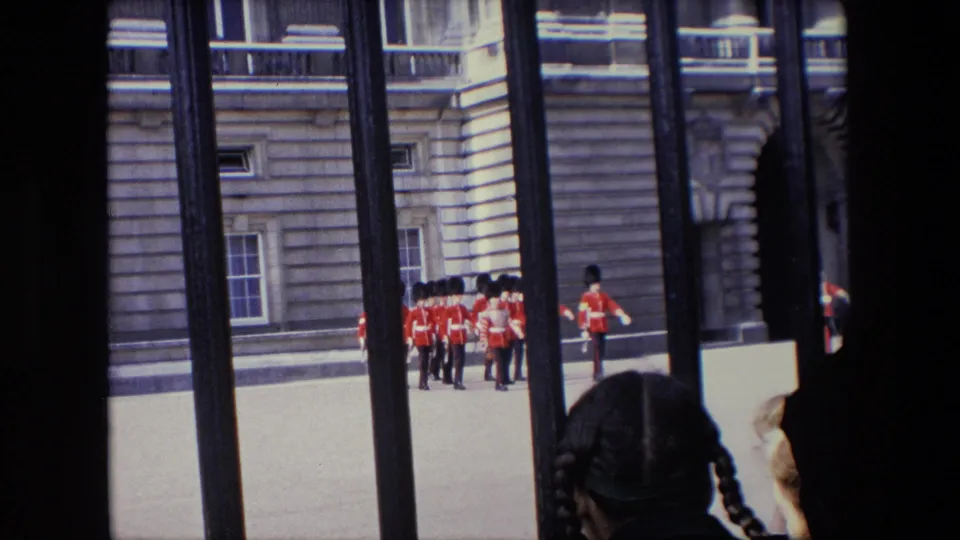 1967 London home-movie frame showing guards marching in red uniforms across a palace courtyard, partially obscured by iron railings and a foreground spectator.