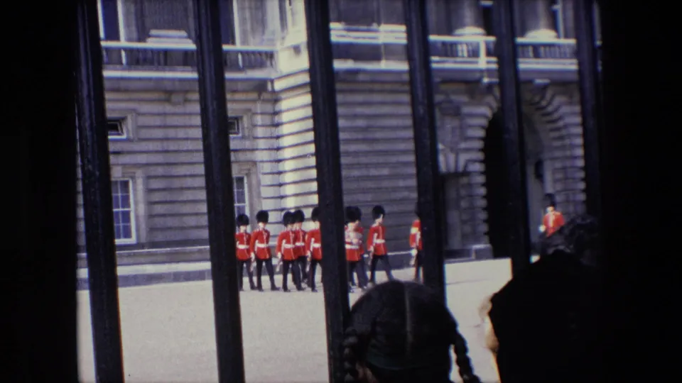1967 London frame with Queen's Guard soldiers moving in formation beside a stone palace facade, viewed from behind railings with partial spectator faces at the edge.