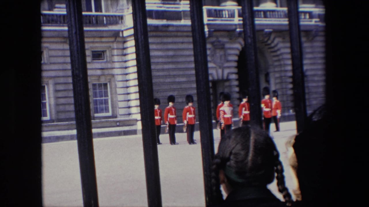 1967 London home-movie frame showing British guards in red tunics and tall black hats seen through iron railings, with a child or young spectator in the foreground.