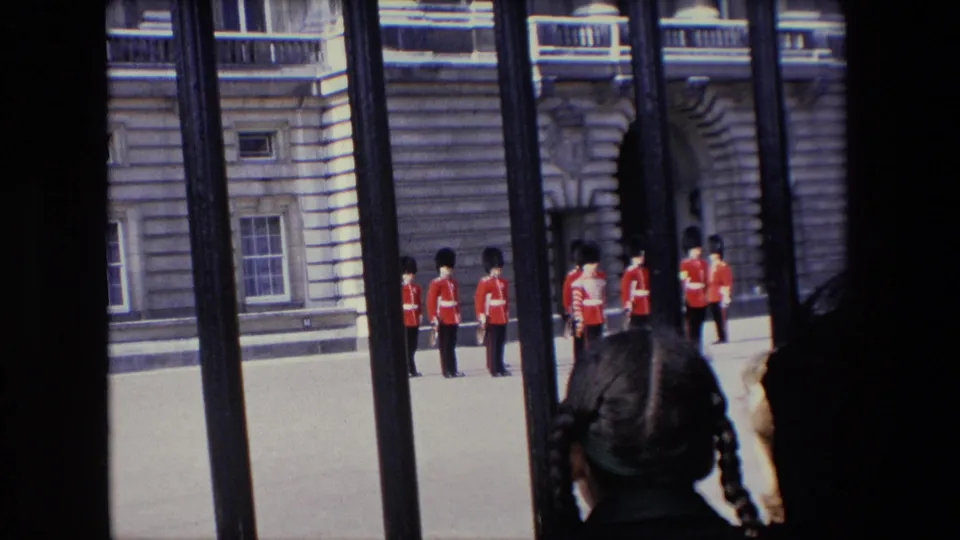 1967 London home-movie frame showing British guards in red tunics and tall black hats seen through iron railings, with a child or young spectator in the foreground.