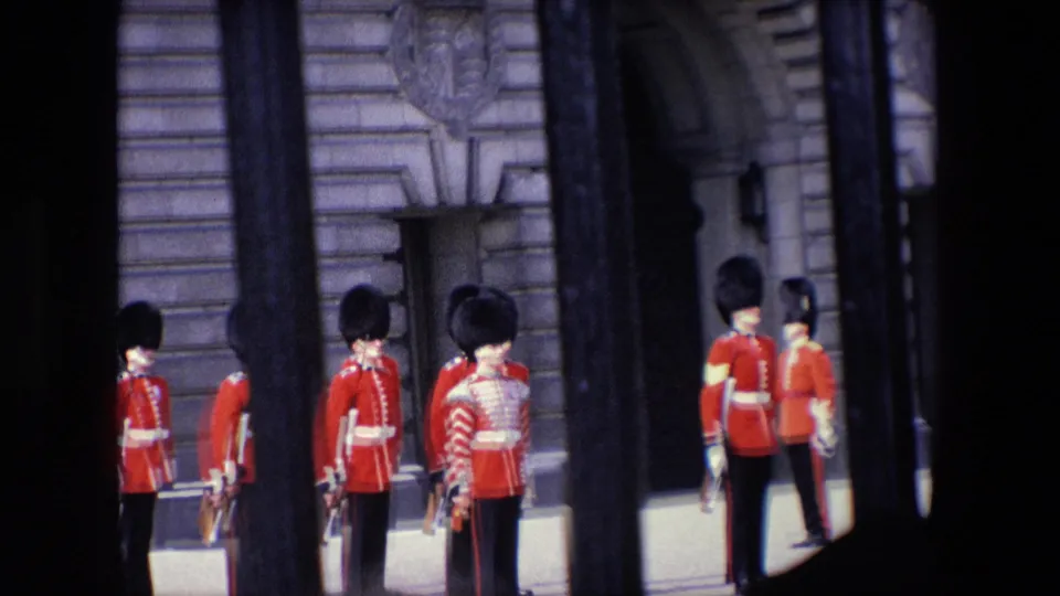 Closer 1967 London home-movie view of British guards in red tunics and tall black hats standing behind railings near an ornate stone entrance.