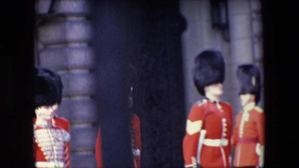 Close 1967 London frame of two British guards in red uniforms and tall black hats, partially blocked by dark foreground railings.