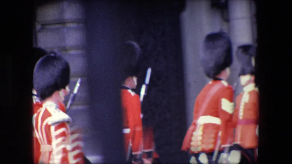 Uniformed red-coated marchers with tall dark hats move across the frame near a dark stone entrance in London, with rifles or staffs visible along the procession.