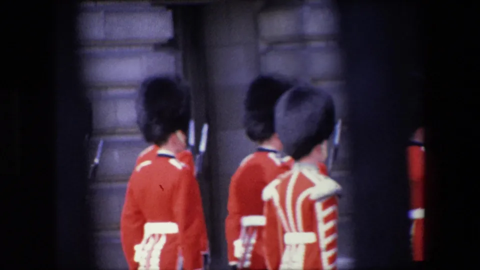 The frame shows several red-coated uniformed marchers from behind, their tall dark hats and white belts visible against stonework and a dark entrance in the 1967 London clip.