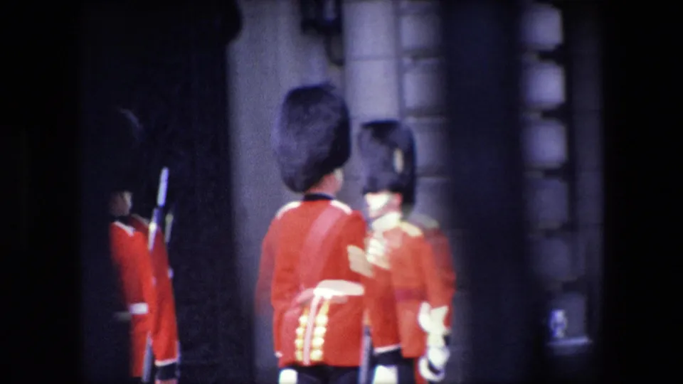 A close, soft 8mm frame shows two red-coated uniformed figures with tall dark hats near a stone doorway in London, with motion blur and strong shadow across the scene.