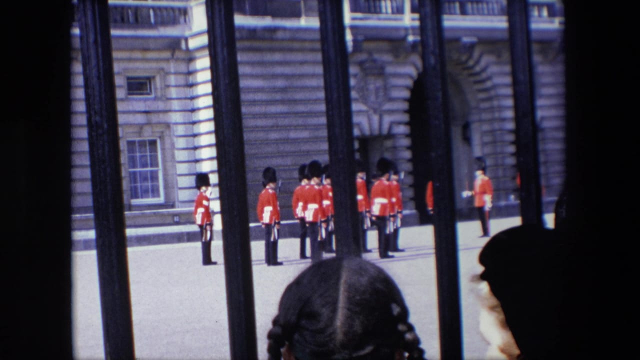 In 1967 London, Royal Guards in red ceremonial uniforms stand at attention in a courtyard, viewed through black railings with tourists partially visible at the bottom of the frame.