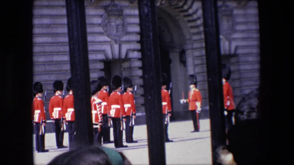 A closer 1967 London home-movie view shows Royal Guards lined up near an arched stone doorway, with black railings and tourist heads interrupting the frame.