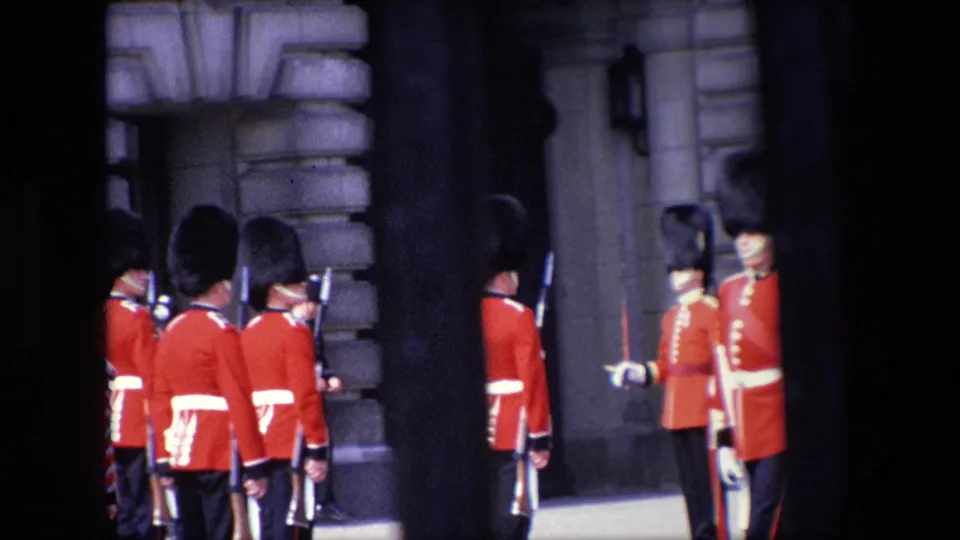 A close visitor-shot frame shows Royal Guards with rifles near a stone doorway in London, with a uniformed guard facing the line and railings crossing the view.
