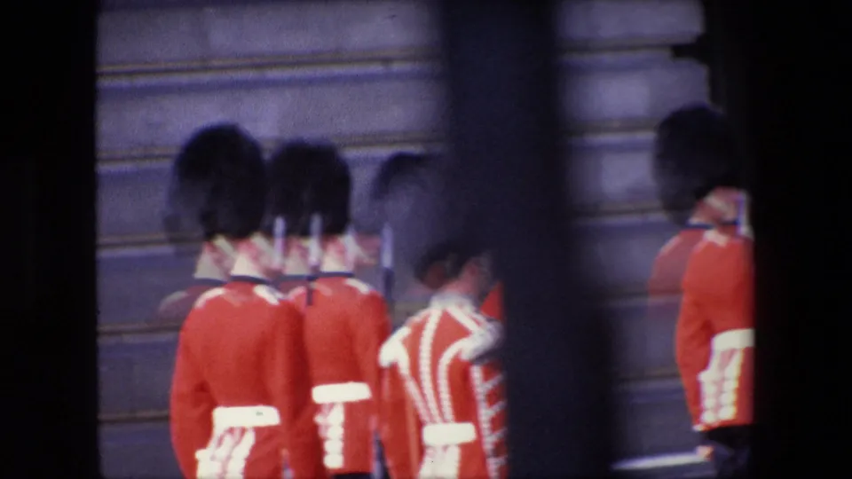 1967 London home-movie frame with multiple ceremonial guards in red uniforms clustered near a stone building, softened by motion blur and foreground obstruction.