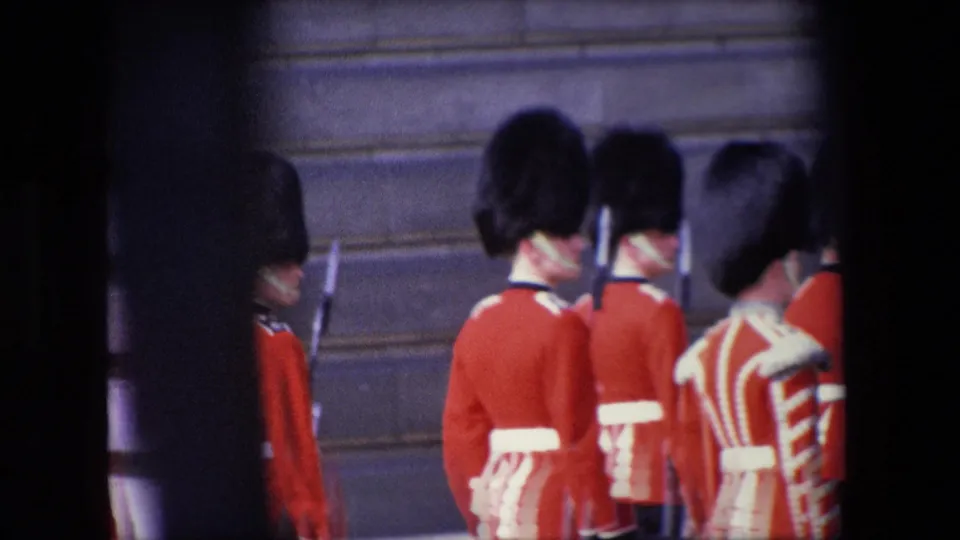 1967 London frame showing several ceremonial guards in red tunics and tall black hats with rifles, viewed from a partially obstructed home-movie angle.