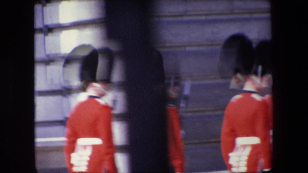 Royal Guards in red uniforms pass a stone building, partly obscured by dark foreground framing.