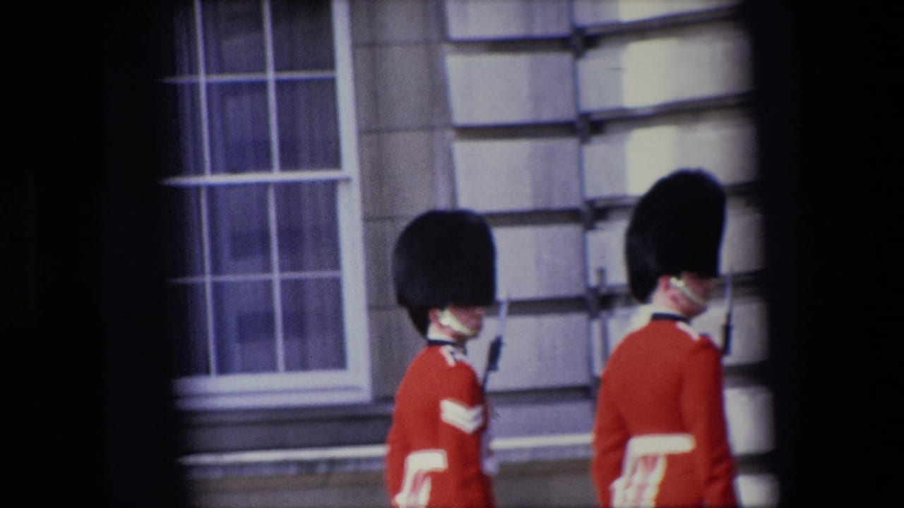 Two Royal Guards in red ceremonial uniforms stand or walk beside a stone facade and window.