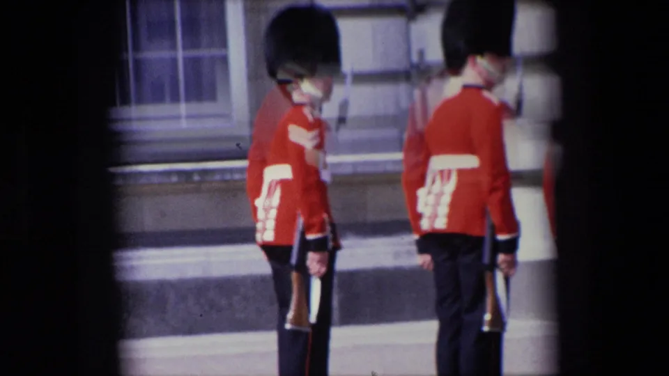 1967 London home-movie frame showing two Royal Guards in ceremonial red uniforms beside steps and a stone facade, with rifles held at their sides.