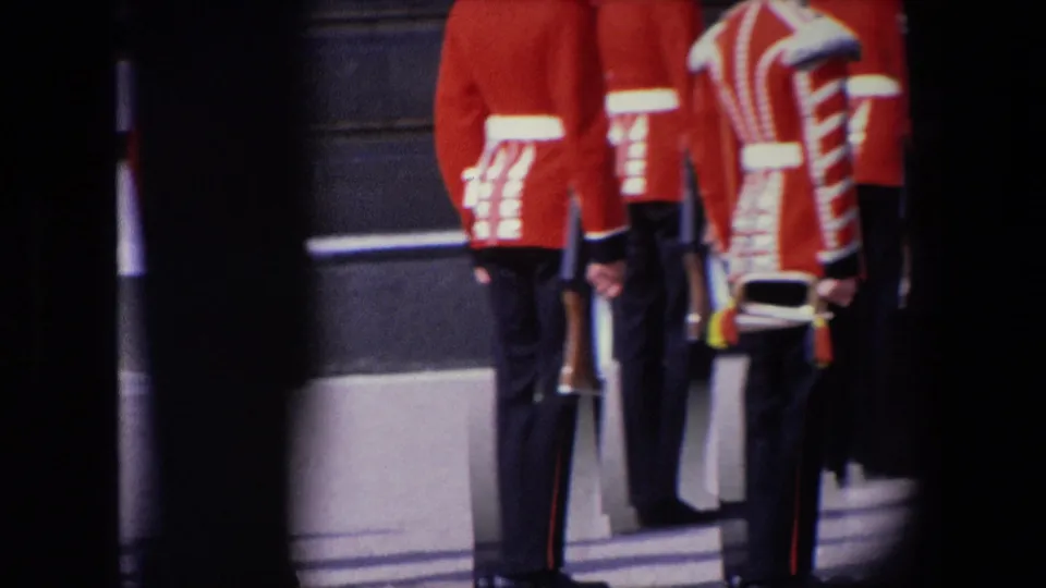 1967 London frame showing several Royal Guards gathered outside a stone building, including red ceremonial coats, tall black headgear, rifles, and a partially visible striped uniform detail.