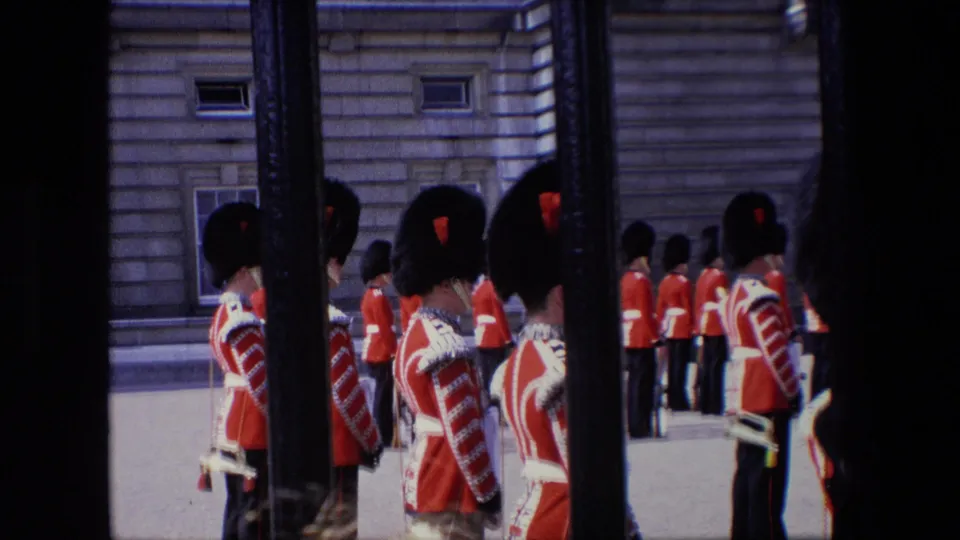 1967 London frame showing red-coated guards in tall dark hats standing at attention near a stone facade, filmed from behind vertical gate bars.