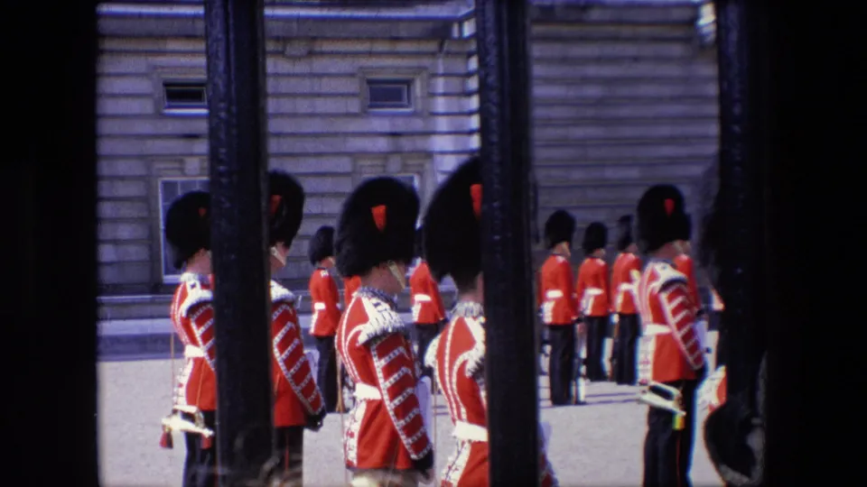 1967 London home-movie view of several red-coated ceremonial guards standing in a line, with black gate bars and a stone exterior framing the scene.