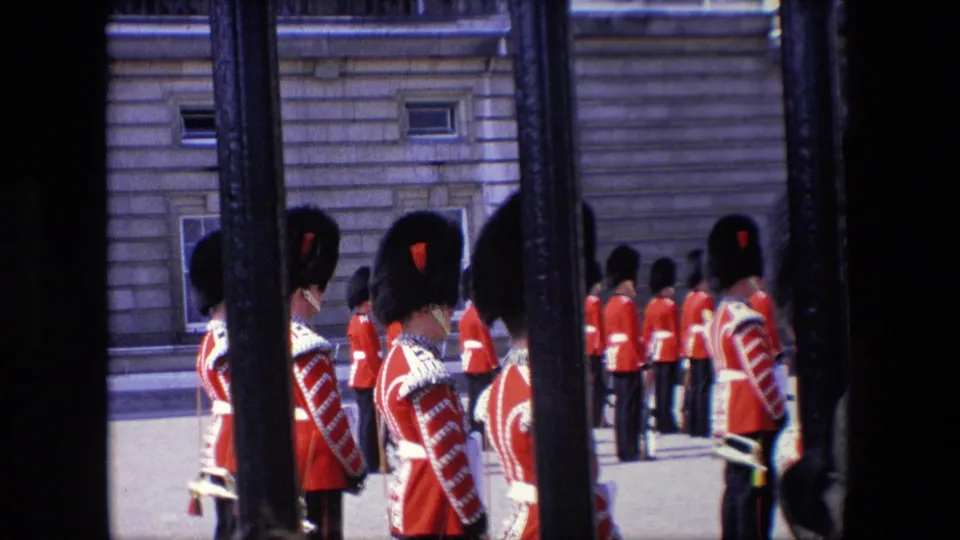 1967 London frame showing ceremonial guards in red uniforms and tall dark hats standing near a stone building, with the camera looking through vertical bars.