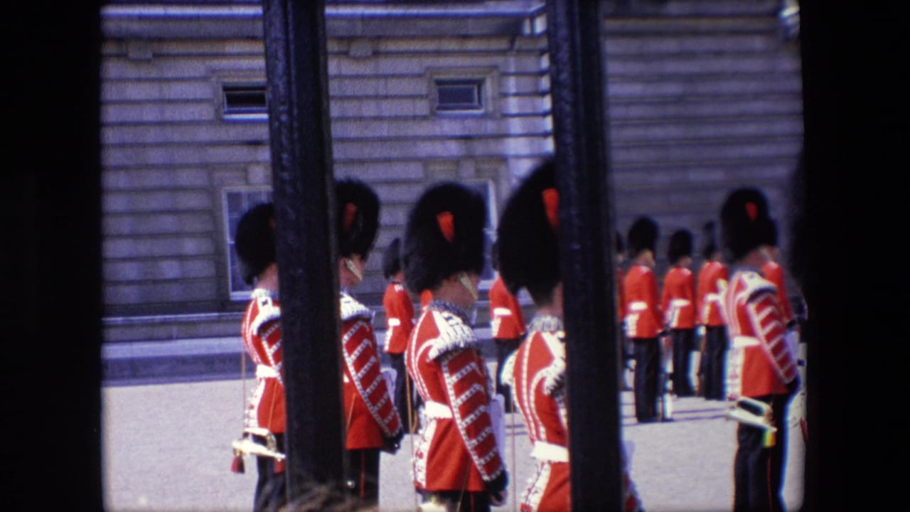 Rows of Royal Guards stand in red uniforms near a stone building, partly obscured by railings.