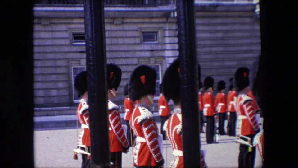 1967 London 8mm frame showing British Royal Guards standing at attention in a courtyard, partially blocked by foreground iron railings.