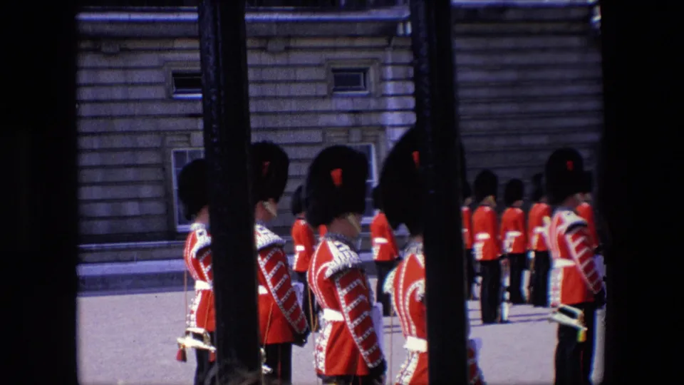 1967 London home-movie frame of Royal Guards in red ceremonial dress standing in rows near a pale stone building, filmed from behind railings.