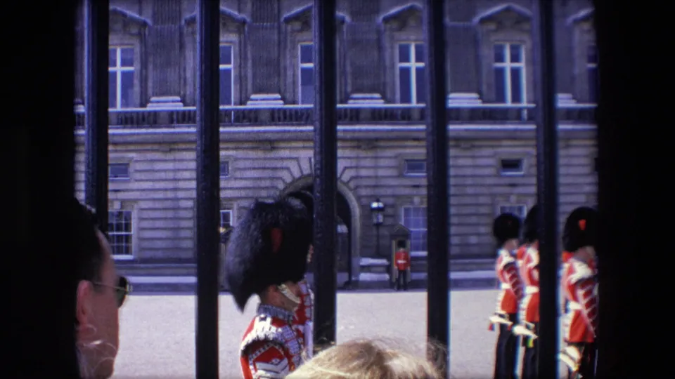 London 1967 8mm frame showing ceremonial guards in red uniforms near railings, with spectators in the foreground and a stone building across the courtyard.