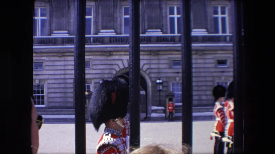 1967 London home-movie frame showing red-uniformed guards and sentry boxes viewed through black railings outside 10 Downing Street.