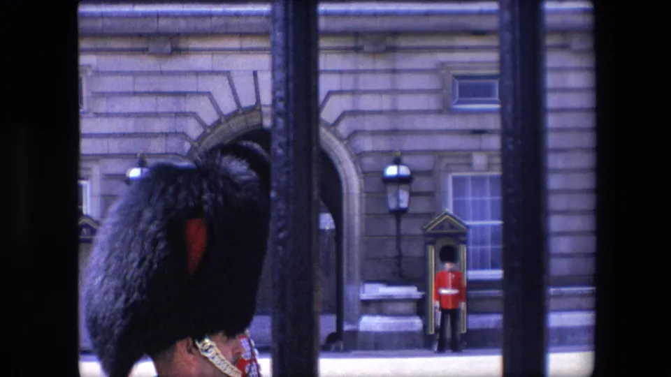 1967 London home-movie frame focused on ceremonial guard headgear in the foreground, with a red-coated sentry by the building behind.