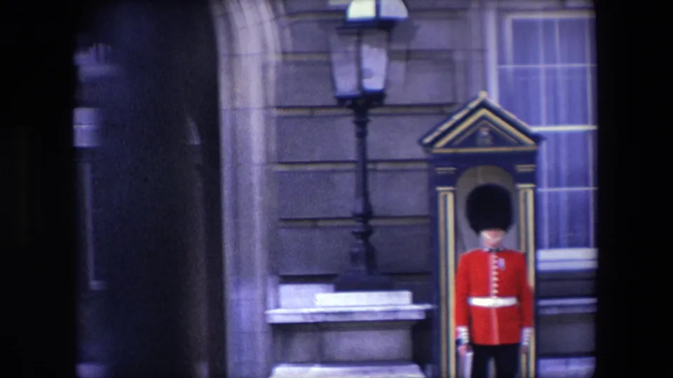 1967 London home-movie frame with a red-coated Royal Guard standing at attention beside a painted sentry box, with a sash window, stone wall, and lamp post nearby.