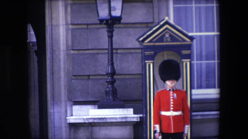 Closer 1967 London home-movie view of the Royal Guard in red uniform and large black hat standing by a sentry box, with a lamp post, stone facade, and window filling the background.