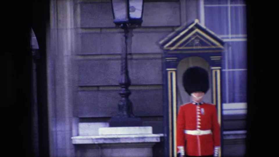 1967 London home-movie frame showing the Royal Guard framed near the right side of the shot, standing at a sentry box beneath a lamp post and in front of a stone building facade.